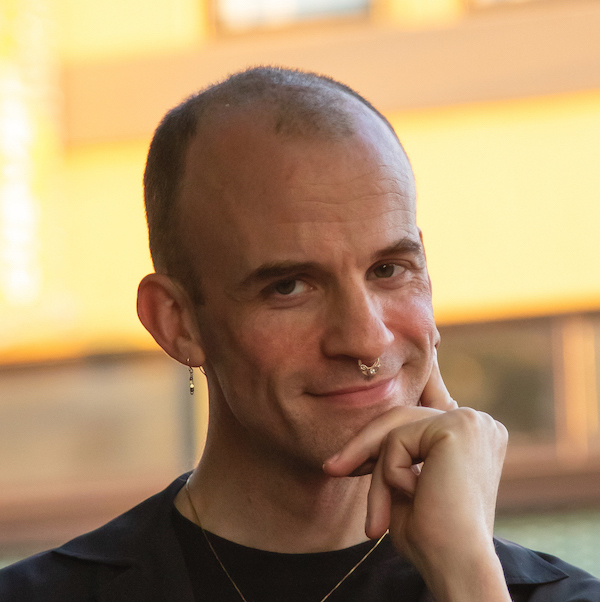 Headshot of Sasha Strong. Sasha is a white person with a shaved head, earrings, a gold nose ring, a necklace, and black dress. They are smiling and looking at the camera.