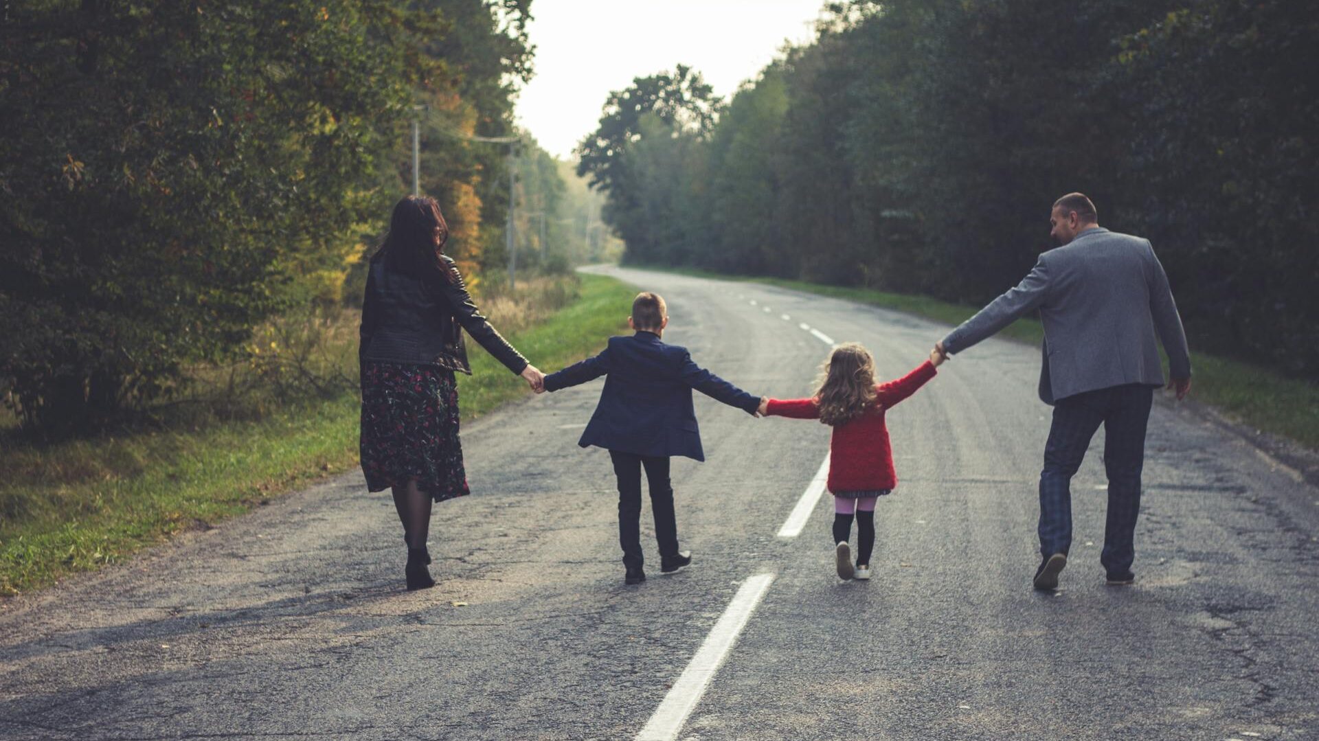 two adults and two children holding hands in a line, walking down a road together